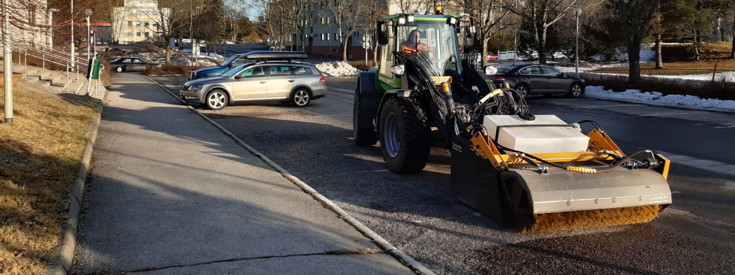 Traktori poistaa hiekoitushiekkaa paikoitusalueelta pyörvällä harjalla.