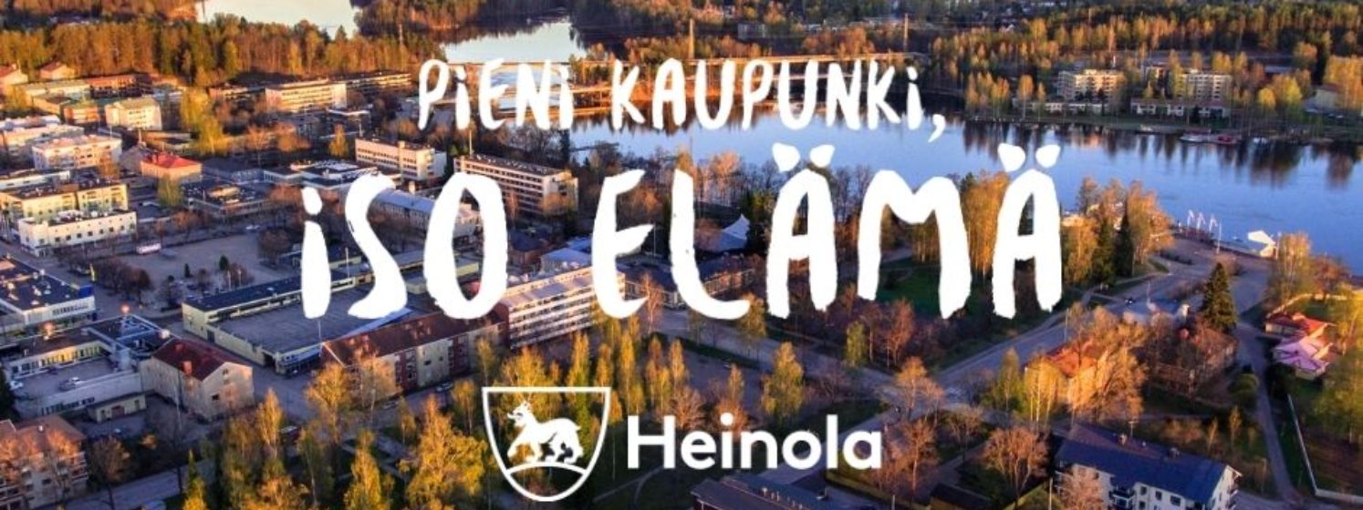 Kuva Heinolan keskustasta auringonlaskun aikaan. Kuvassa iso logo.