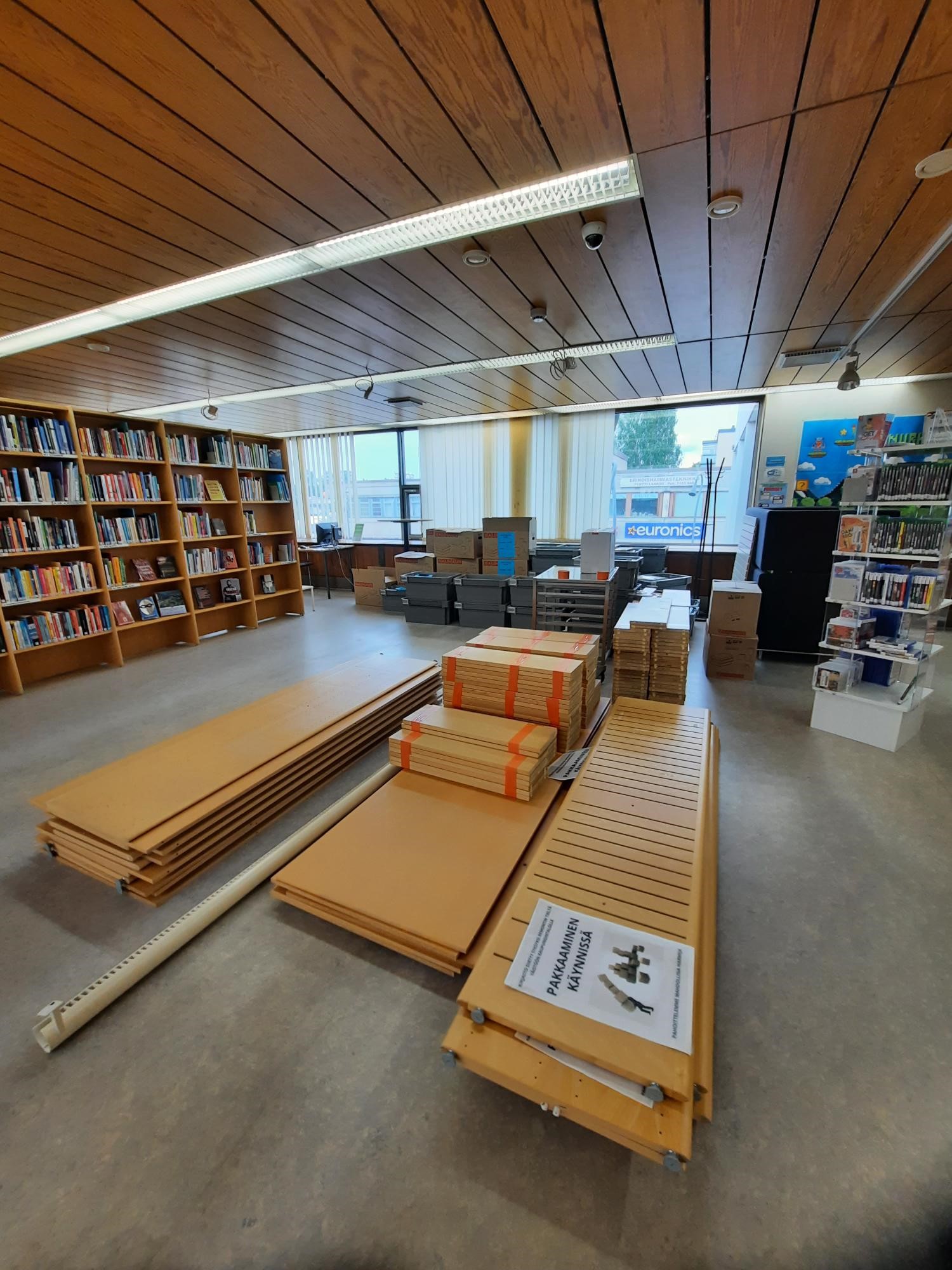 Kirjaston kirjahyllyjä purettuna pinoissa, muuttlaatikoita, taustalla täysiä kirjahyllyjä.